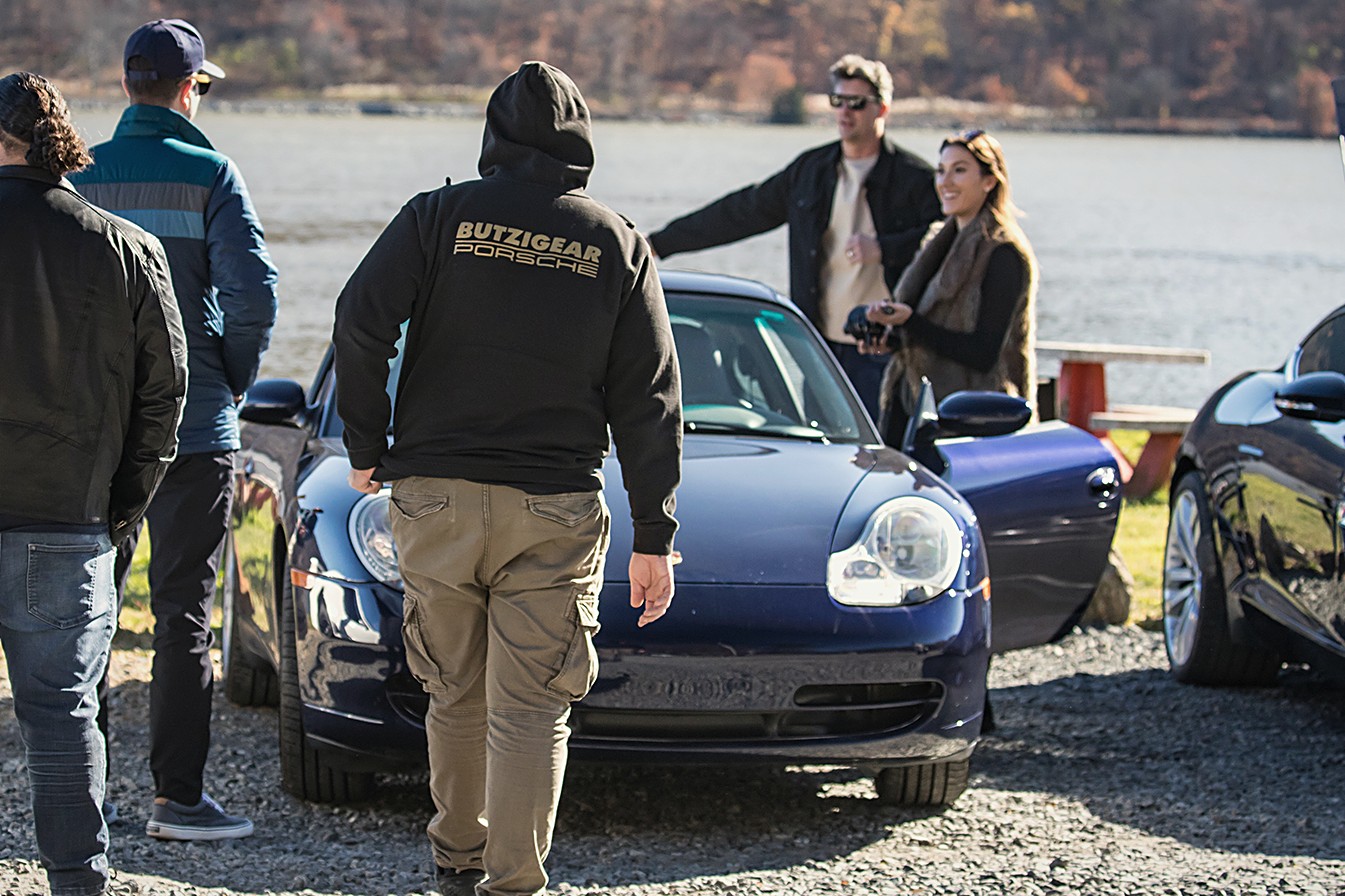 Group talking beside a blue Porsche at a riverside stop, autumn hills behind