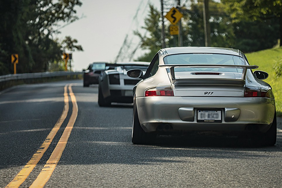 Silver Porsche 911 leading two cars along a double-yellow country road in summer
