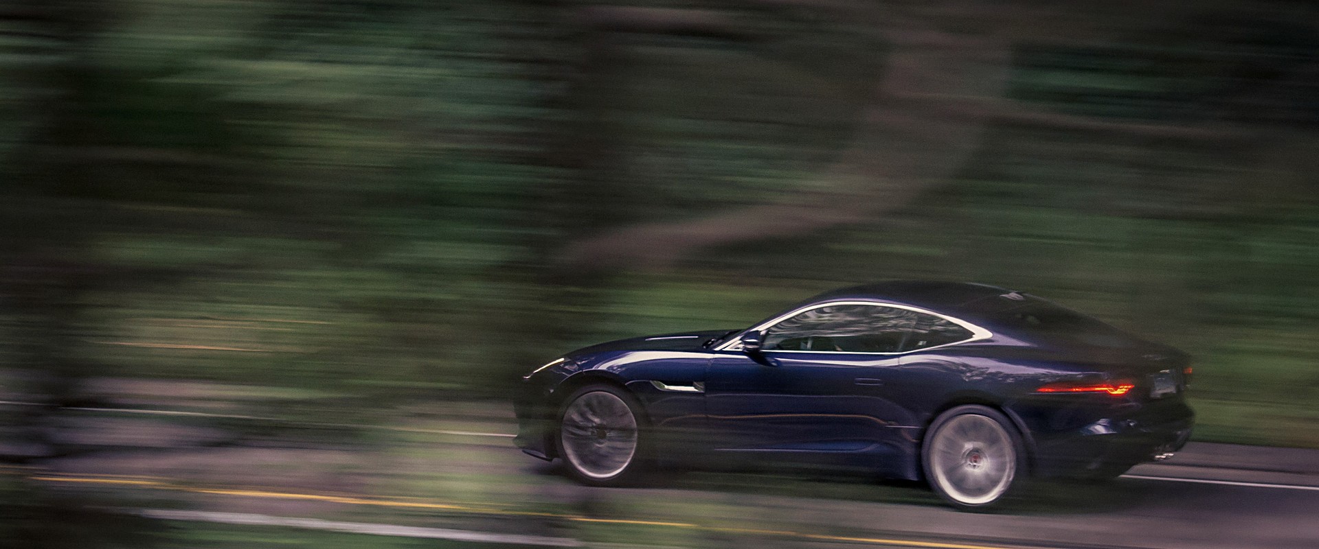 Blue Jaguar F-Type at speed on a tree-lined road, background blurred with motion